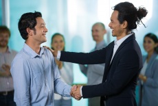 Optimistic business colleagues talking while shaking hands and looking at each other. Happy boss congratulating new employee. People clapping in background. Business event concept